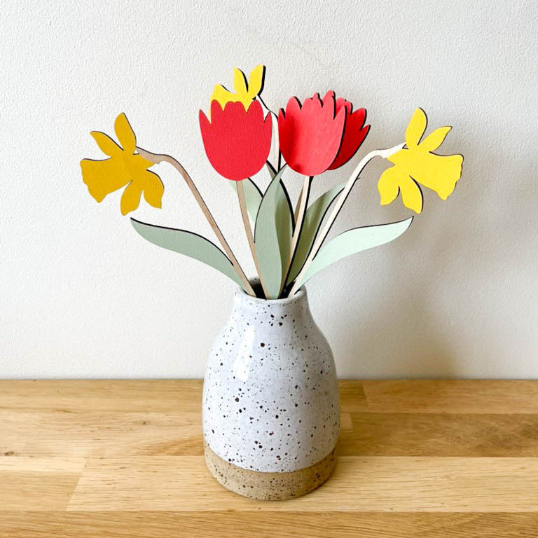 A ceramic speckled vase on a wooden surface against a plain wall, holding wooden flowers in red tulips, yellow daffodils, and green leaves, with bare wood stems.