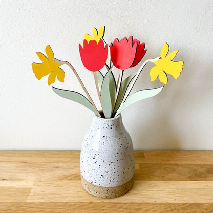 A ceramic speckled vase on a wooden surface against a plain wall, holding wooden flowers in red tulips, yellow daffodils, and green leaves, with bare wood stems.
