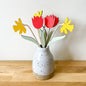 A ceramic speckled vase on a wooden surface against a plain wall, holding wooden flowers in red tulips, yellow daffodils, and green leaves, with bare wood stems.