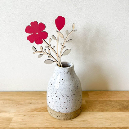 A small speckled ceramic vase sits on a light wood tabletop against a white wall. A stem of decorative wooden flowers in red with bare wooden stems is placed in the vase.