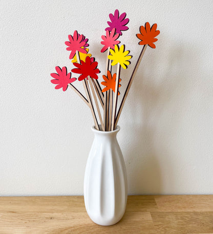 A white ceramic vase on a light wood surface against a white wall, holding several wooden flower stems with brightly coloured flowers painted in pink, red, orange and yellow.