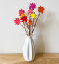 A white ceramic vase on a light wood surface against a white wall, holding several wooden flower stems with brightly coloured flowers painted in pink, red, orange and yellow.