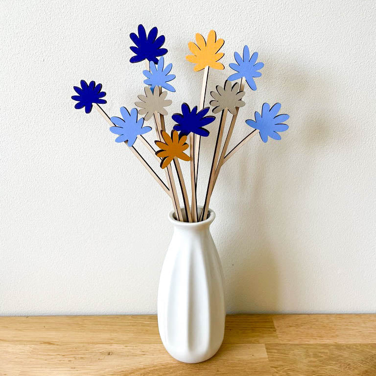 A white ceramic vase on a light wood surface against a white wall, holding a bouquet of wooden flower stems in shades of blue, yellow, and grey.