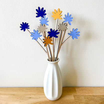 A white ceramic vase on a light wood surface against a white wall, holding a bouquet of wooden flower stems in shades of blue, yellow, and grey.