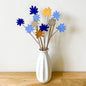 A white ceramic vase on a light wood surface against a white wall, holding a bouquet of wooden flower stems in shades of blue, yellow, and grey.