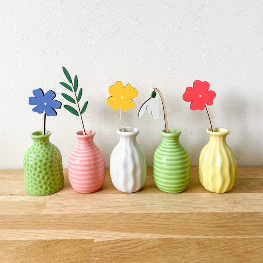 Five small decorative ceramic vases in green, pink, white, light green, and yellow, lined up on a light wood surface against a plain off-white wall. Each vase holds a wooden flower stem with a different color flower: blue, yellow, white, red, and yellow.