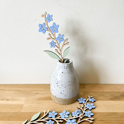 A decorative wooden floral branch with small blue flowers and pale green leaves, displayed in a speckled ceramic vase on a light wood surface against a plain off-white wall.